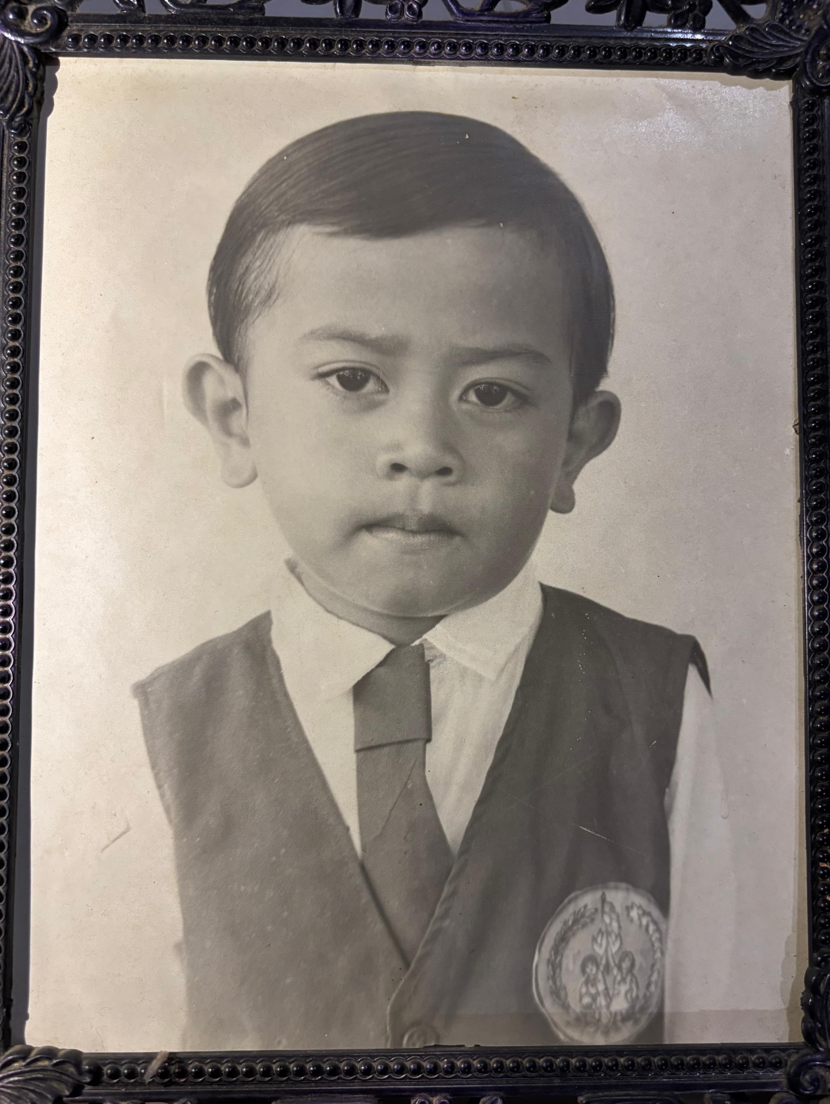 Potret hitam-putih Gugum Ridho Putra kecil berseragam sekolah — kemeja putih, dasi, dan rompi berlambang — di dalam bingkai foto berukir.